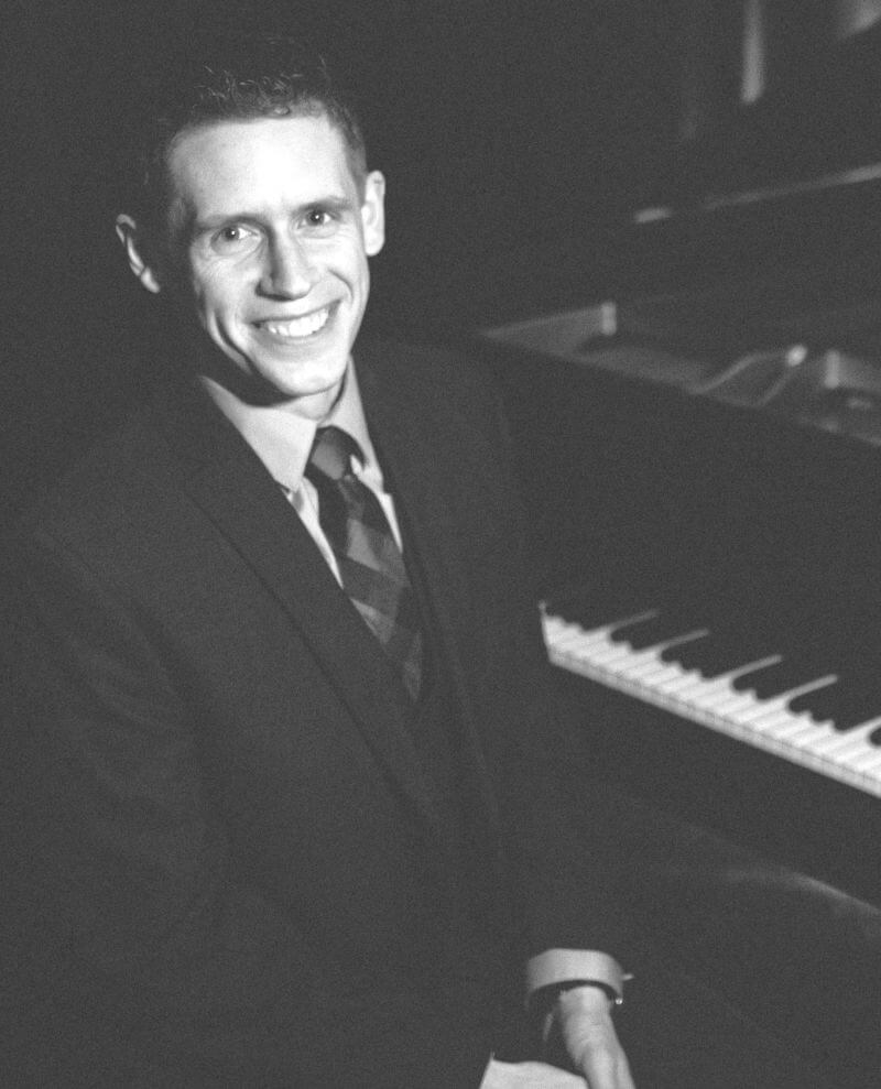 Pianist and composer Jay Frost in B&W in front of piano on stage